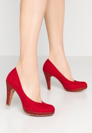 Women's legs wearing red suede high heels with glittery heels and platform soles on a light floor and background.