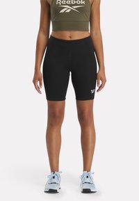 Bermudas negras de bicicleta hechas de un tejido elástico, con un diseño ajustado y el logo de Reebok en el dobladillo izquierdo. Combinadas con un crop top verde oliva.