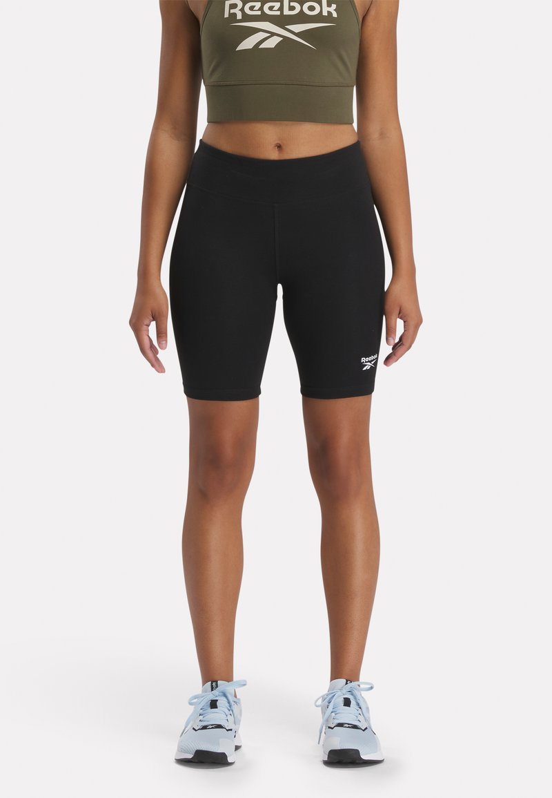 Bermudas negras de bicicleta hechas de un tejido elástico, con un diseño ajustado y el logo de Reebok en el dobladillo izquierdo. Combinadas con un crop top verde oliva.