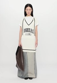 White knit tunic with black accents and graphic text, layered over a light gray and denim maxi skirt. Model holds a brown tote bag.