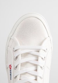 Sneaker blanc avec un dessus texturé et brillant, bout arrondi, lacets blancs, œillets en argent et accent de logo rouge sur le côté.