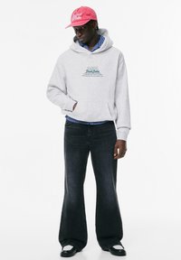 Felpa grigia con cappuccio e dettagli testuali verdi, sovrapposta a una maglietta a righe blu e bianche. I jeans neri a zampa completano il look insieme a un cappellino rosa.
