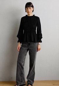 Blusa de peplum negra con fruncidos, de manga larga y puños con volantes, combinada con jeans de pierna ancha grises. El modelo está posando frente a un fondo neutro.