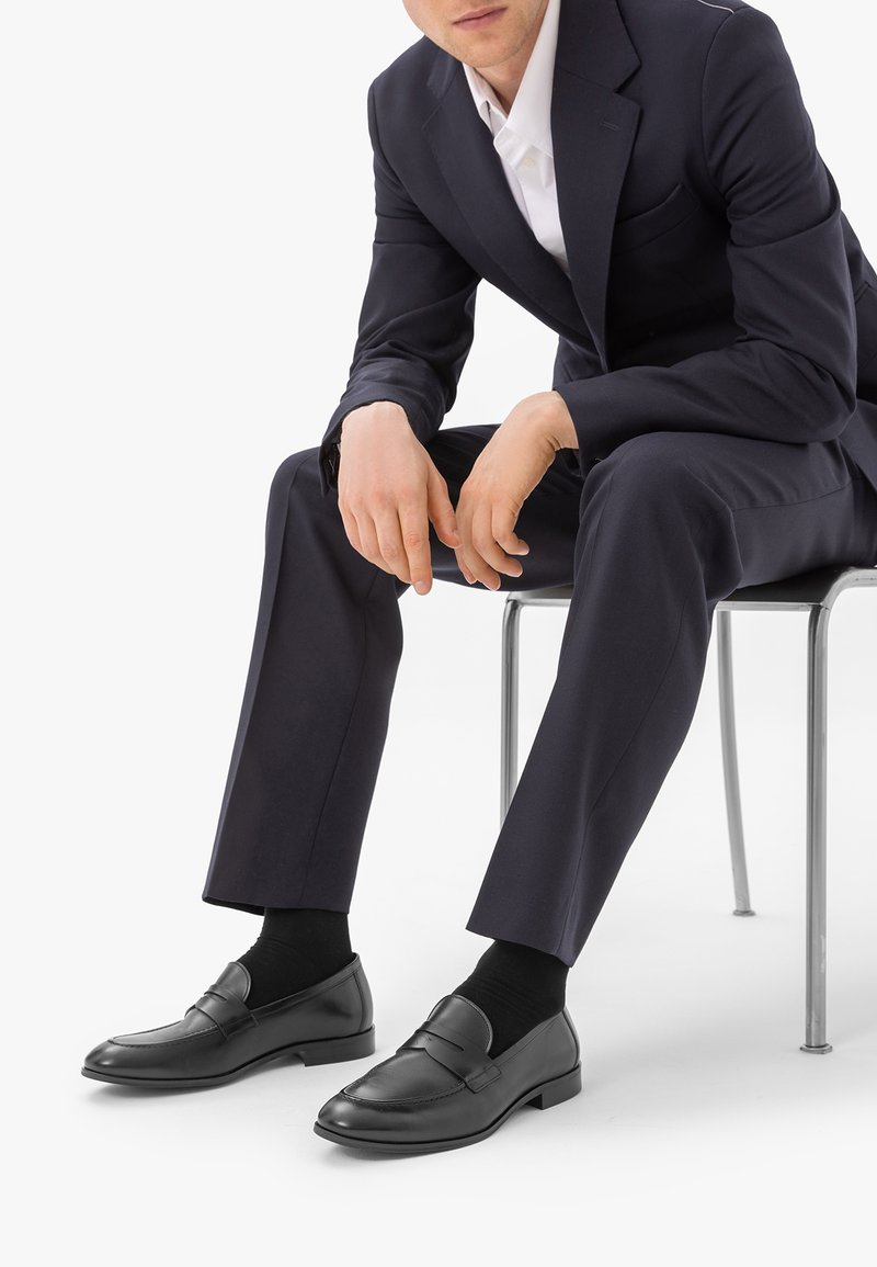 Homme assis sur une chaise portant un costume noir, une chemise blanche, des chaussettes noires et des mocassins en cuir noir, les mains reposant entre les genoux.