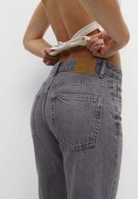Jean en denim gris à taille haute, présentant une étiquette en cuir marron carré à l'arrière et un tissu texturé. La personne est en train de nouer une sangle.