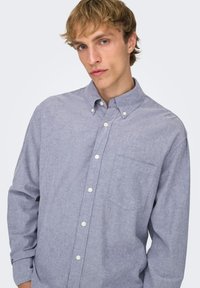 Lyseblå skjorte med knapper, krave, lange ærmer, enkelt brystlomme og tekstureret stof med et diskret mønster.