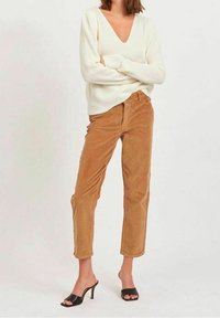 Suéter de punto acanalado color crema con escote en V combinado con pantalones de pana color beige. El modelo lleva unas sandalias de tacón abierto negras. Diseño sencillo y moderno.