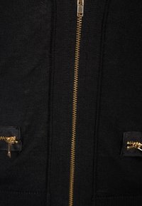 Svart stickad zip-up-jacka med gyllene dragkedja mitt fram och två fickor med dragkedjor. Texturen är mjuk med ribbade detaljer.