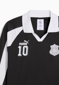 Αθλητική φανέλα Puma σε ασπρόμαυρο με τον αριθμό 10 στο μπροστινό μέρος και ένα περίπλοκο έμβλημα στο δεξί στήθος, με άσπρο γιακά.
