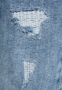 Světle modrá denímová látka s dvěma roztrhanými záplaty, které odhalují bílou tkaninu pod nimi.