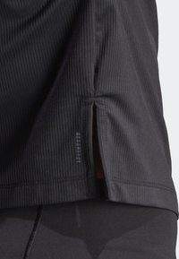 Top atlético negro con textura acanalada, aberturas laterales y una etiqueta de logo visible. Diseñado para flexibilidad y transpirabilidad.