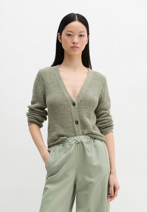 Kobieta o długich czarnych włosach, ubrana w jasnozielony dziany kardigan na guziki oraz luźne spodnie ze ściągaczem, stojąca na jednolitym tle.