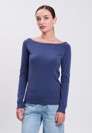 Femme à la peau claire et aux yeux bleus portant un pull bleu marine à manches longues avec col bateau et un jean bleu clair, debout devant un fond uni.