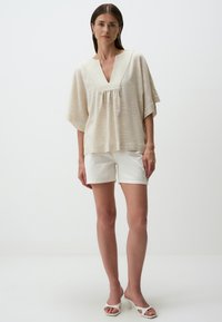 Blusa beige a maniche corte con scollo a V, realizzata in tessuto leggero, con finitura testurizzata e dettagli plissettati, abbinata a pantaloni corti bianchi e sandali.
