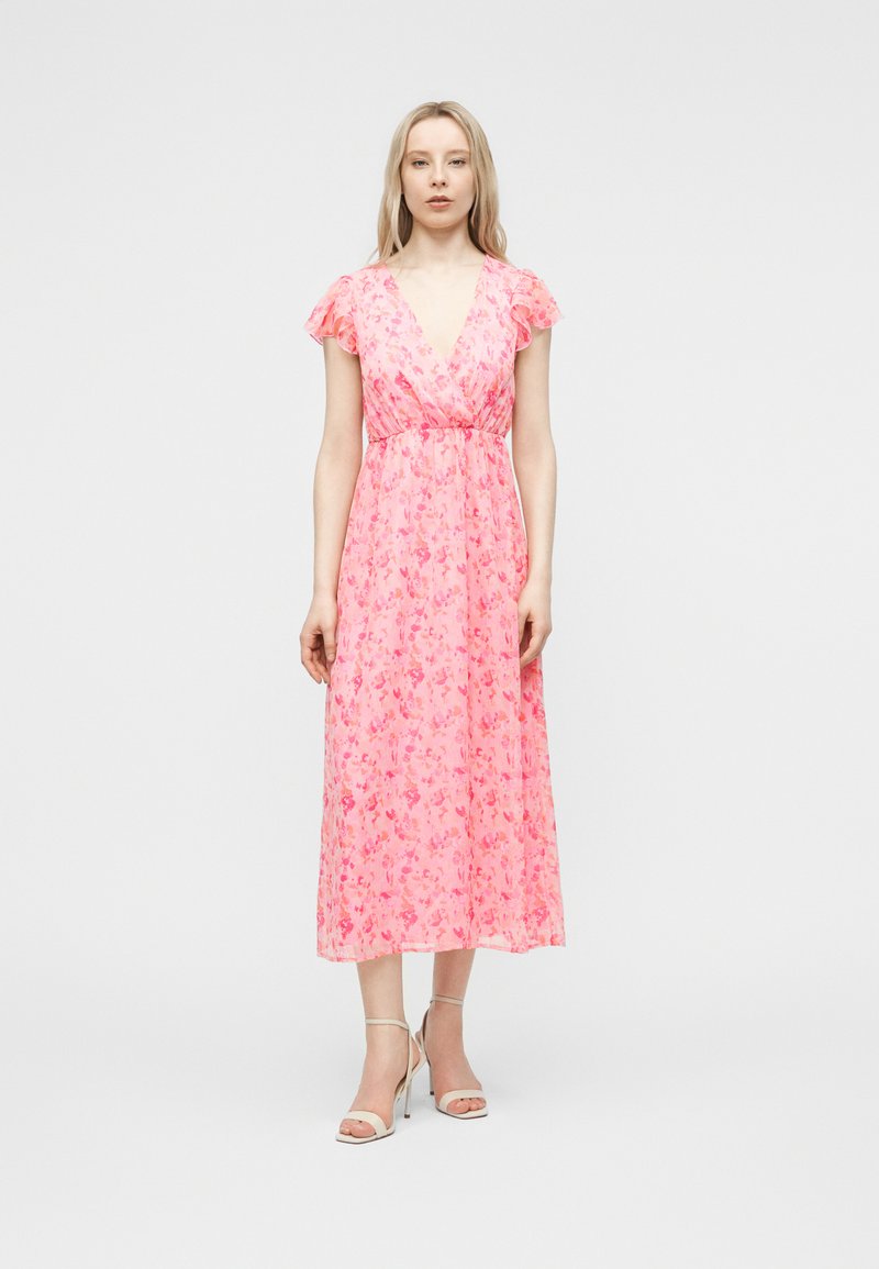 Γυναίκα που φοράει ροζ μίντι φόρεμα με λουλούδια, με φαρδιά μανίκια και βαθύ V-ντεκολτέ, συνδυασμένο με λευκά σανδάλια με ψηλό τακούνι, όρθια.