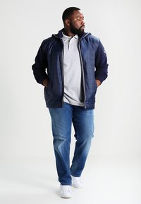 Chaqueta de plumón azul marino con cremallera, camiseta tipo polo gris, vaqueros azules y zapatillas blancas. La chaqueta tiene mangas texturizadas y bolsillos laterales.