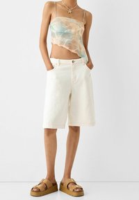 Shorts in denim bianco con vestibilità comoda e orli risvoltati, abbinati a una canottiera trasparente con fantasia tie-dye in tonalità pastello. Sandali platform marroni con dettaglio fibbia.