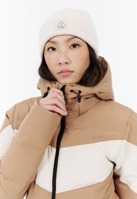 Hnedo-biely pruhovaný puffer kabát s vysokým golierom, čiernym zipsom a nastaviteľnou kapucňou, nosený s krémovou pletenou čiapkou s logom.