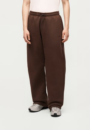 Osoba ubrana w luźne brązowe spodnie dresowe ze ściągaczem w talii oraz jasnoszare buty sportowe na jednolitym tle.