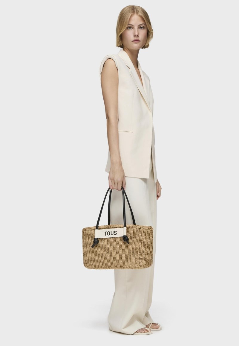 Bolso tote de paja trenzada con asas negras, que presenta un acento de cuero blanco adornado con "TOUS" en la parte frontal. Forma rectangular.