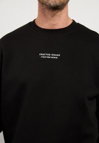 Felpa nera con testo bianco "CRAFTED GOODS - PIER ONE DENIM" sul petto, indossata da una persona con barba chiara e collo visibile.