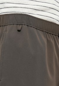 Γκρι ελαστική μέση με κορδόνι, με απαλή, λεία ύφανση και διακριτική γυαλάδα. Ριγέ τελείωμα πουκάμισου ορατό πάνω από το παντελόνι.