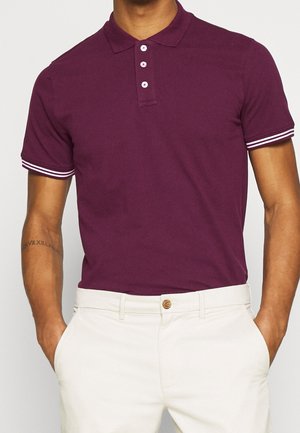 Man wearing a short-sleeve, dark purple polo shirt with white stripes on cuffs and white pants, hands in pockets.