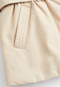 Beżowa tkanina z kieszenią z wyhaftowanymi detalami, miękka w dotyku, z delikatnymi marszczeniami w talii dla dodatkowego kształtu.