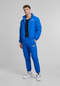 Blue lightweight tracksuit with elastic waistband and cuffs, featuring a logo on the jacket and pants. Worn with white sneakers.