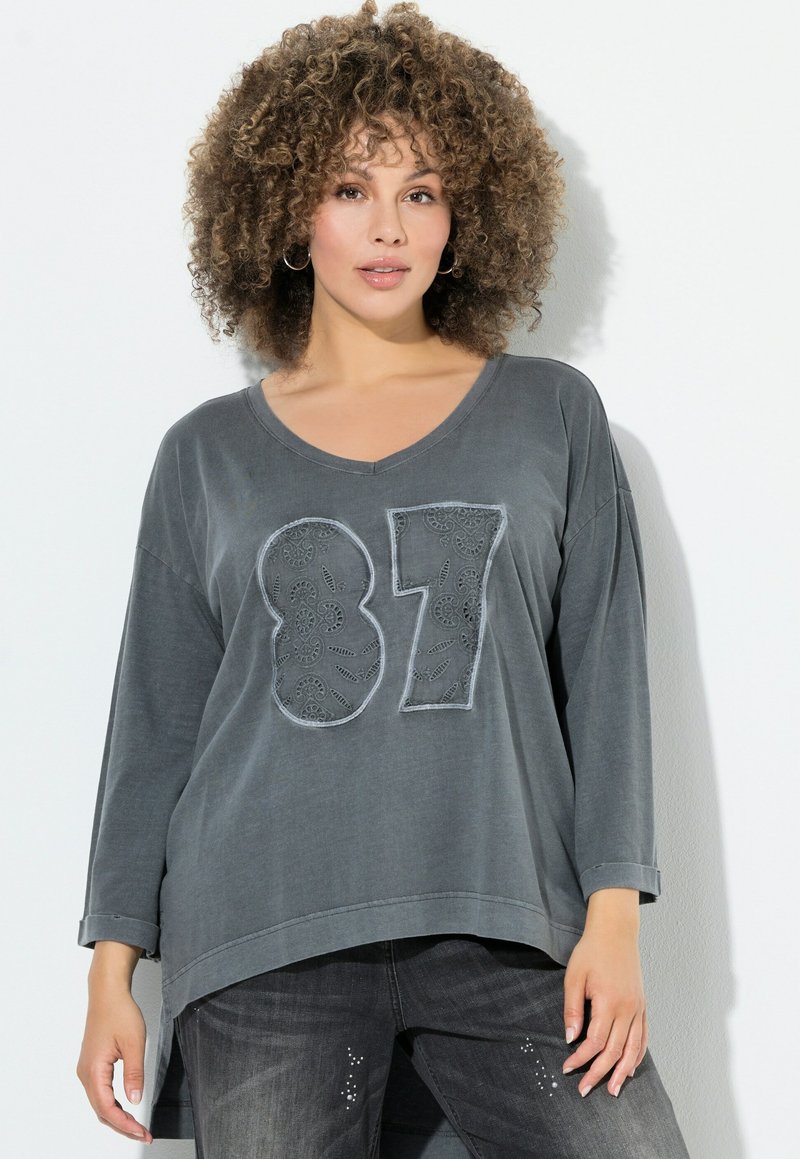 Graues Langarmshirt mit V-Ausschnitt, versehen mit großen, gestickten Zahlen "87" aus kontrastierendem Stoff. Weicher Stoff und lockere Passform.