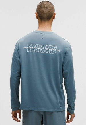 Langärmeliges Trainingsshirt aus hellblauem Stoff. Es verfügt über einen Rundhalsausschnitt, nahtlose Schultern und weißen Text auf der Rückseite: „TRAINING“.