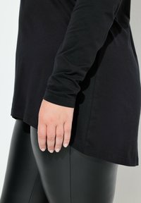 Czarny top z długim rękawem z zaokrąglonym dołem, zestawiony z dopasowanymi czarnymi legginsami. Gładka tekstura i minimalistyczny design bez widocznych zdobień.