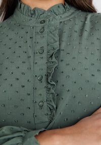 Zbli�żenie na zieloną bluzkę o strukturalnej fakturze z małymi wypukłymi kropeczkami, z przodu z zapięciem na guziki oraz falbaniastym wykończeniem przy dekoldzie i mankiecie rękawa.