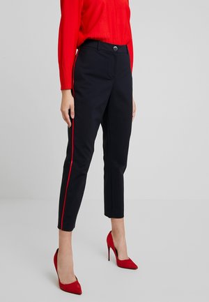 Kvinde iført sorte slim-fit bukser med røde sidelister, rød sweater og røde højhælede sko, stående på en ensfarvet grå baggrund.