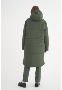 Grön vadderad lång kappa med huva, med en slät textur och horisontell sömnad. Matchad med gröna byxor och svarta skor.