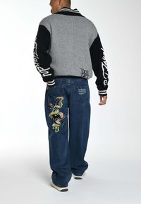 Maglione a maglia grigio e nero con ampio colletto, caratterizzato da intricati dettagli bianchi. Jeans blu con design ricamato colorato sulla gamba sinistra.