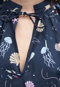 Μπλούζα σε ναυτικό μπλε με βαθύ V λαιμόκοψη δεμένη στον γιακά, διακοσμημένη με παστέλ θαλάσσια πλάσματα όπως μέδουσες, ψάρια, αστερίες και κοχύλια.