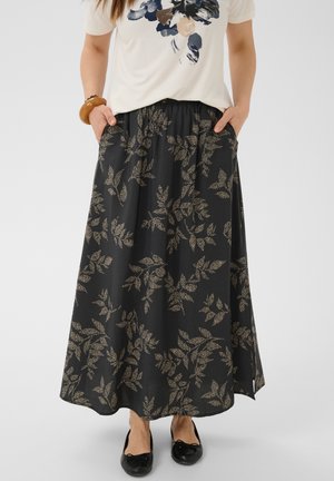 Femme portant une jupe longue noire avec des motifs de feuilles beiges, un t-shirt blanc à imprimé floral, des ballerines noires et un bracelet en bois, les mains dans les poches.