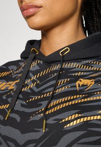 Moletão preto com padrões abstratos em dourado e cinza. Apresenta cordões com pontas douradas e um logo no peito. O material parece texturizado.
