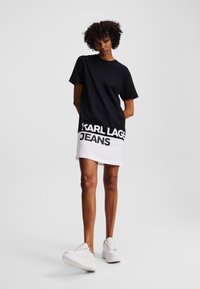 Juodas T-marškinukų suknelė su balta apdaila, ant kurios įspaustas ryškus baltas tekstas "KARL LAGERFELD JEANS". Lygus medvilnės audinys, laisvas fasonas, trumpomis rankovėmis.