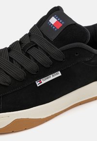 Sorte semskede sneakers med teksturerte svarte lisser, hvit og gummisåle, og et Tommy Jeans logo-patch med røde, hvite og blå farger.