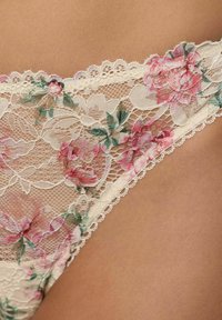Crème kantondergoed met roze en groene bloemen borduursels op een gebruinde huid, waarbij de gedetailleerde stoftextuur en het patroon zichtbaar zijn.