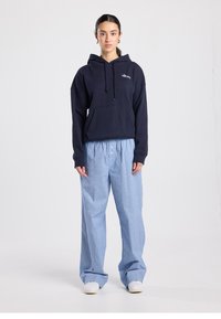 Sweat-shirt à capuche bleu marine avec poche avant, associé à un pantalon large à rayures bleu clair. Le mannequin porte des baskets blanches.