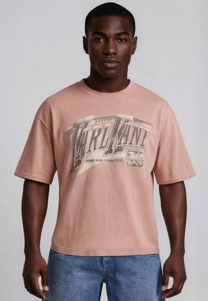 Joven hombre negro que lleva una camiseta rosa clara con el texto “MARLMANI” y vaqueros azules, de pie frente a un fondo gris liso.