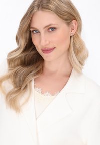 Hellbeige Wolle Blazer mit Reverskragen über einem cremfarbenen Oberteil mit Spitze, mit langen, lockigen Haaren und dezentem Make-up. Klare blaue Augen.