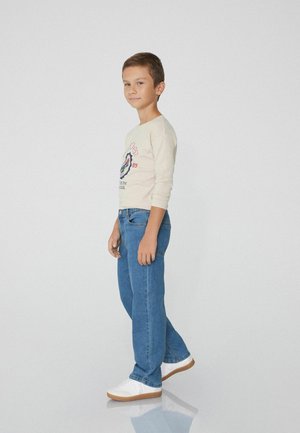 Helle beige Langarmshirt mit grafischem Druck, kombiniert mit blauen Jeans und weißen Sneakers mit braunen Sohlen. Einfaches Design, lässiger Schnitt.