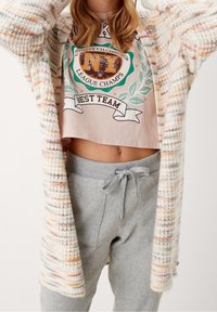 Grober Strickcardigan mit mehrfarbigen Sprenkeln, kombiniert mit einem kurzen Grafik-T-Shirt mit einem Abzeichen-Design und grauen Jogginghosen mit Kordelzug.