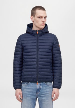 Un homme porte une veste matelassée bleu marine à capuche, dotée de tirettes de fermeture éclair orange, avec un jean bleu clair, sur un fond uni.