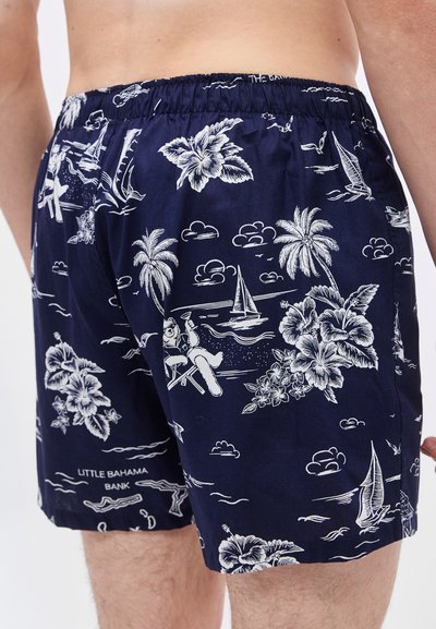 Μπλε ναυτικό μαγιό με λευκό τροπικό σχέδιο που απεικονίζει φοίνικες, λουλούδια, ιστιοφόρα και μια φιγούρα που χαλαρώνει κρατώντας ένα ποτό.