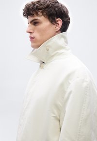 Νέος άνδρας με σγουρά μαλλιά, που φοράει παλτό με ψηλή γιακά σε ανοιχτό μπεζ χρώμα, κοιτάζει πλαγίως μπροστά σε απλό λευκό φόντο.